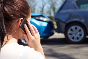 woman on cell phone calling insurance company while looking at her crashed blue car.