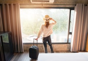 woman with hat on and holding onto suitcase looking out of a large window